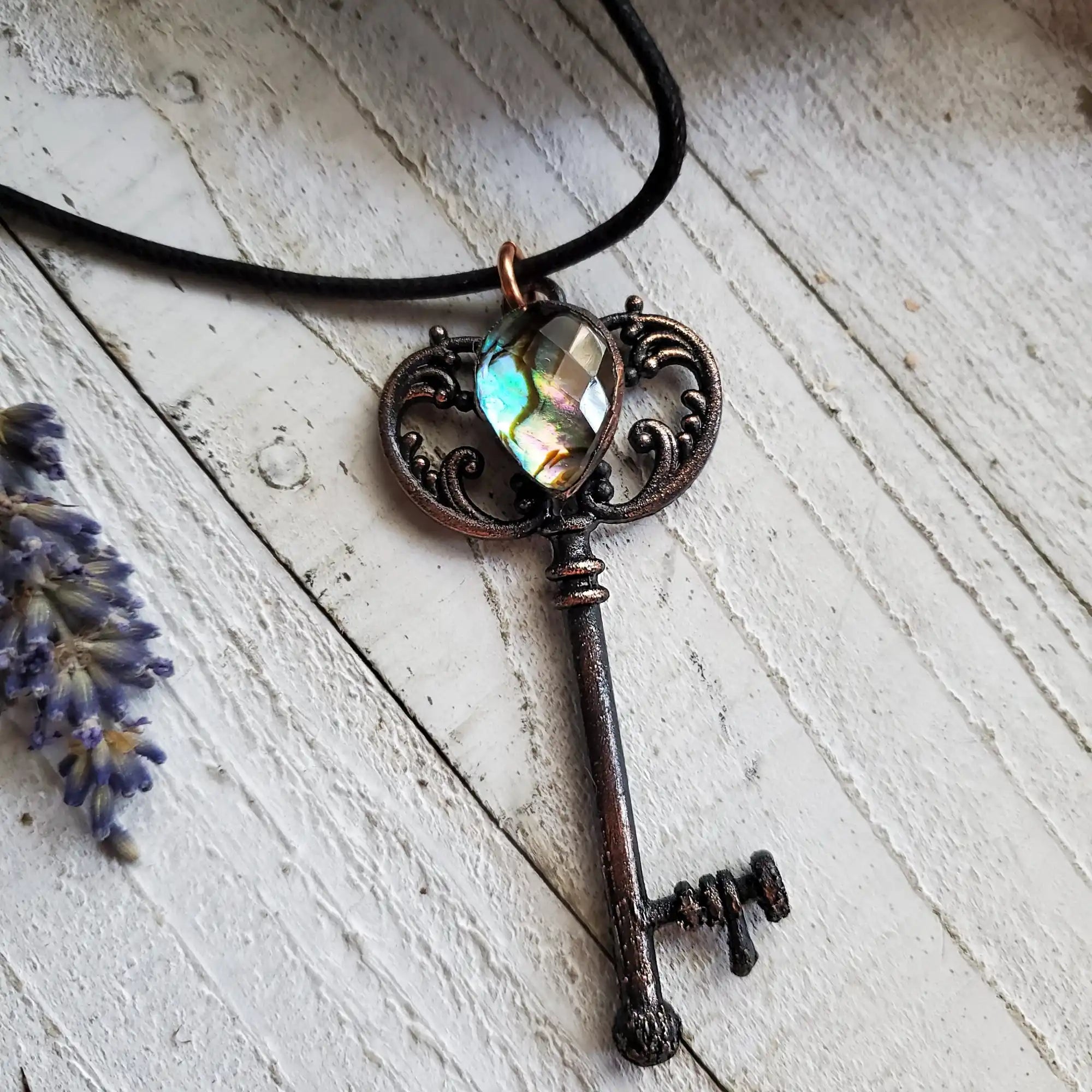 An antique-style key pendant with an iridescent heart-shaped gemstone, on a black cord necklace.