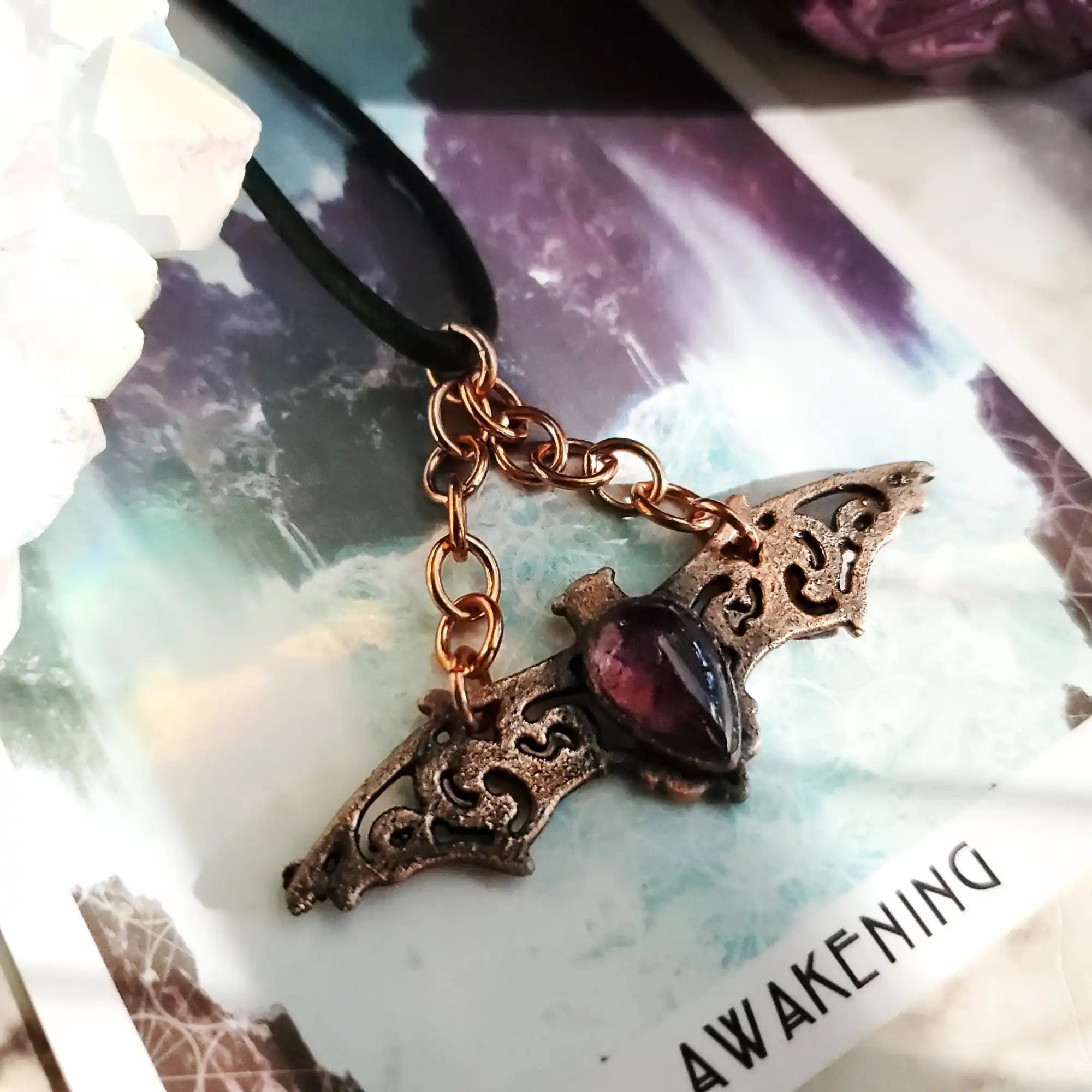 A bronze-colored bat-shaped necklace pendant with an ornate, filigree design and a dark red, teardrop-shaped gem in the center, suspended from a black cord by copper-colored chains.