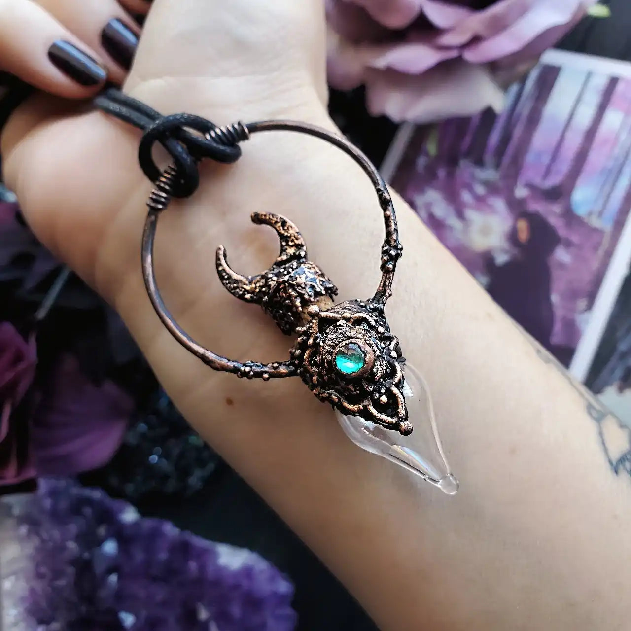 A bronze-toned, horned skull pendant with an iridescent blue-green gem and a clear, pointed crystal dangling from a black cord.