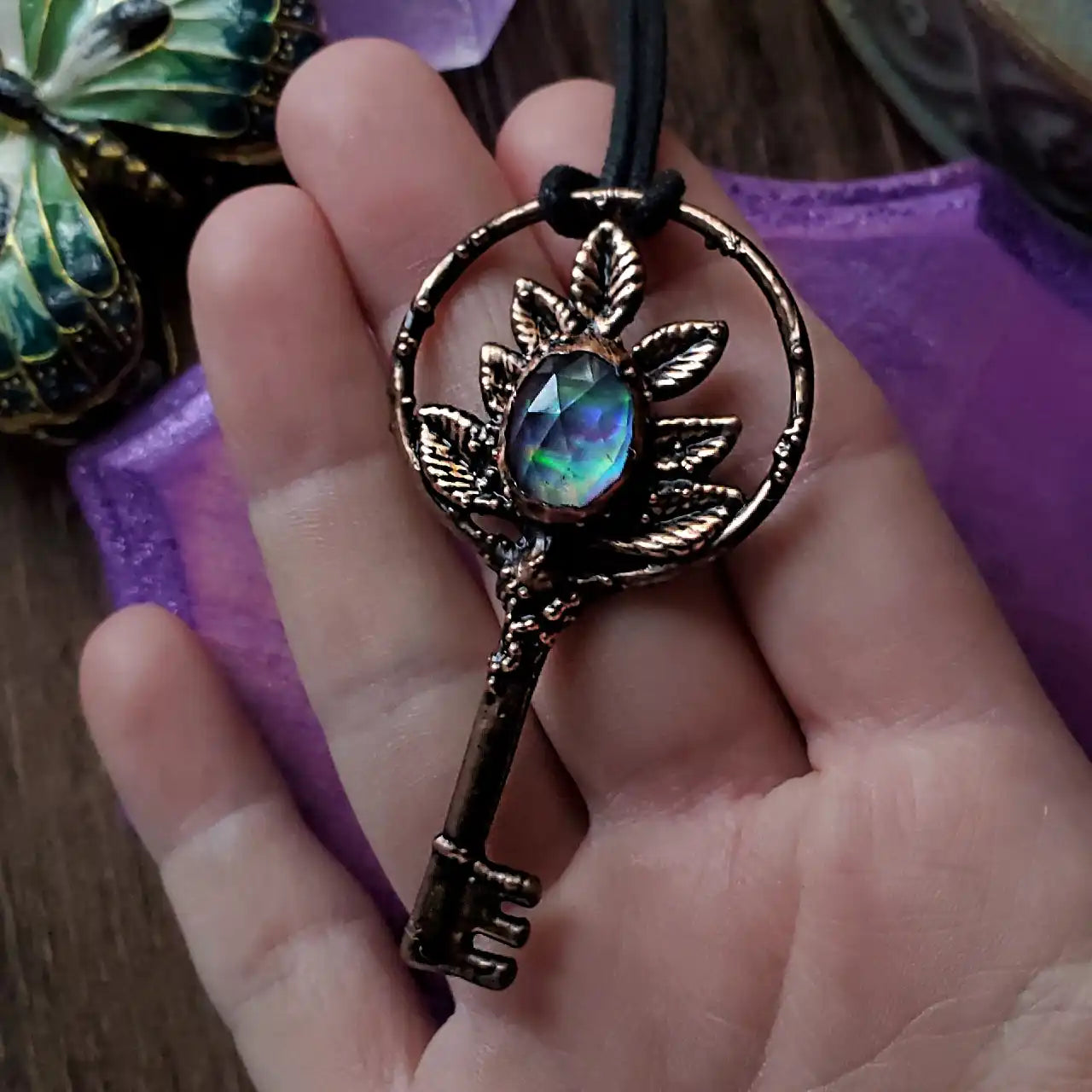 Bronze-toned key pendant with a vibrant iridescent oval gem and leaf accents.