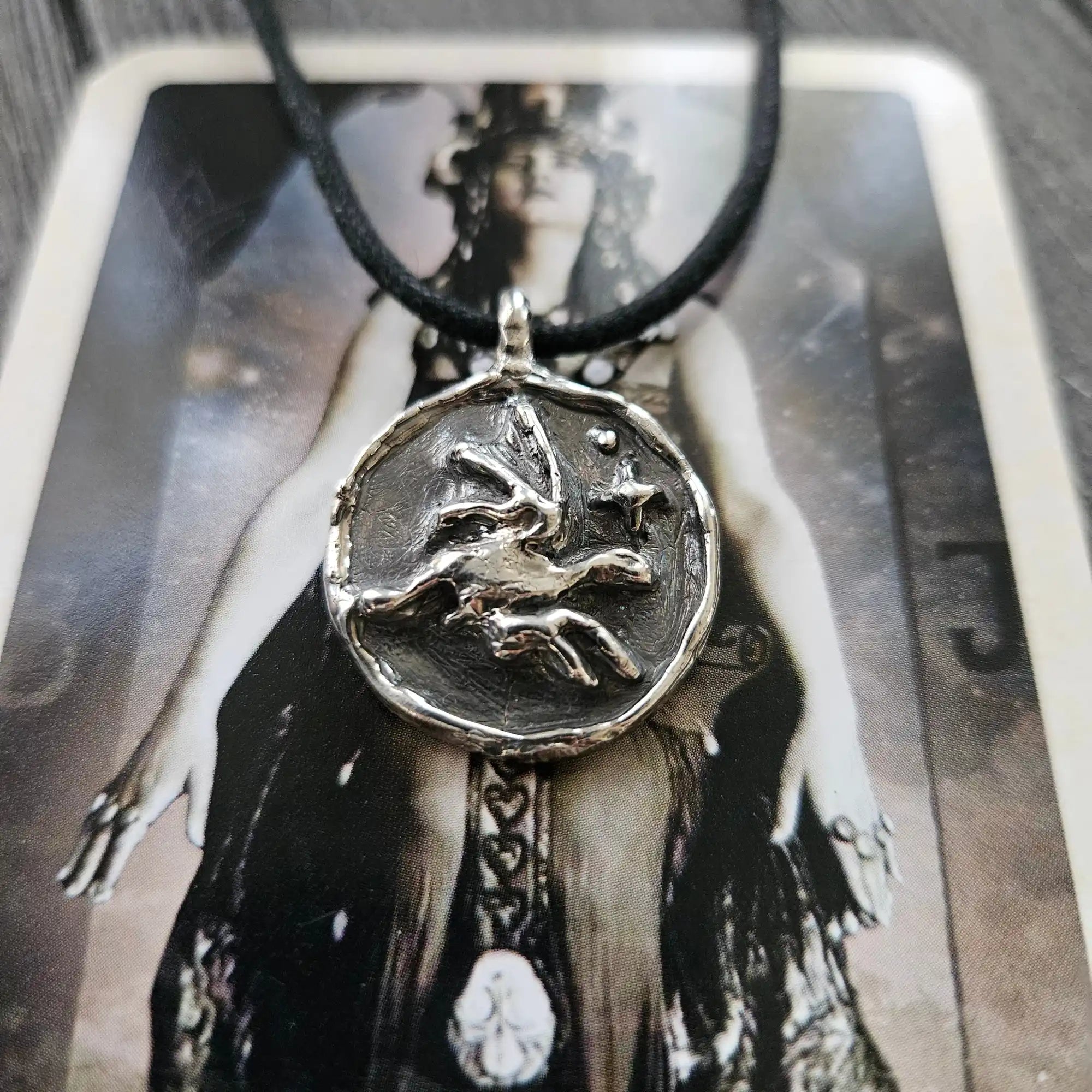A circular silver pendant featuring a winged creature and stars, on a black cord necklace.