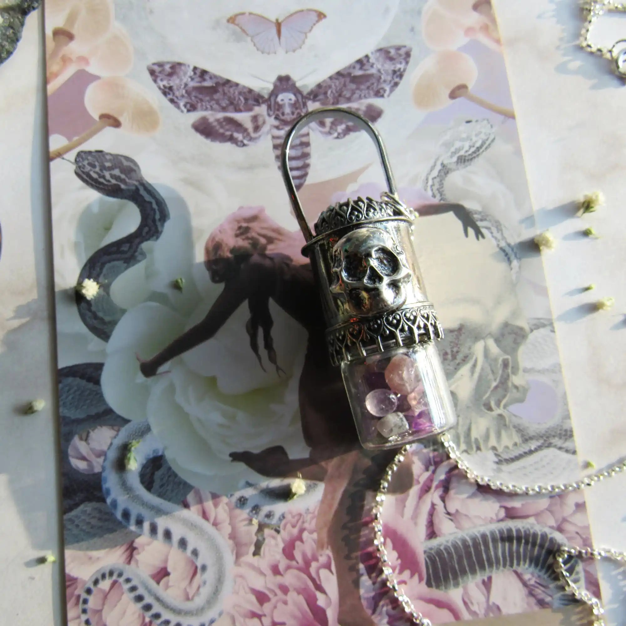 A clear glass vial with a silver skull and ornate metalwork, containing small pink and clear crystals.