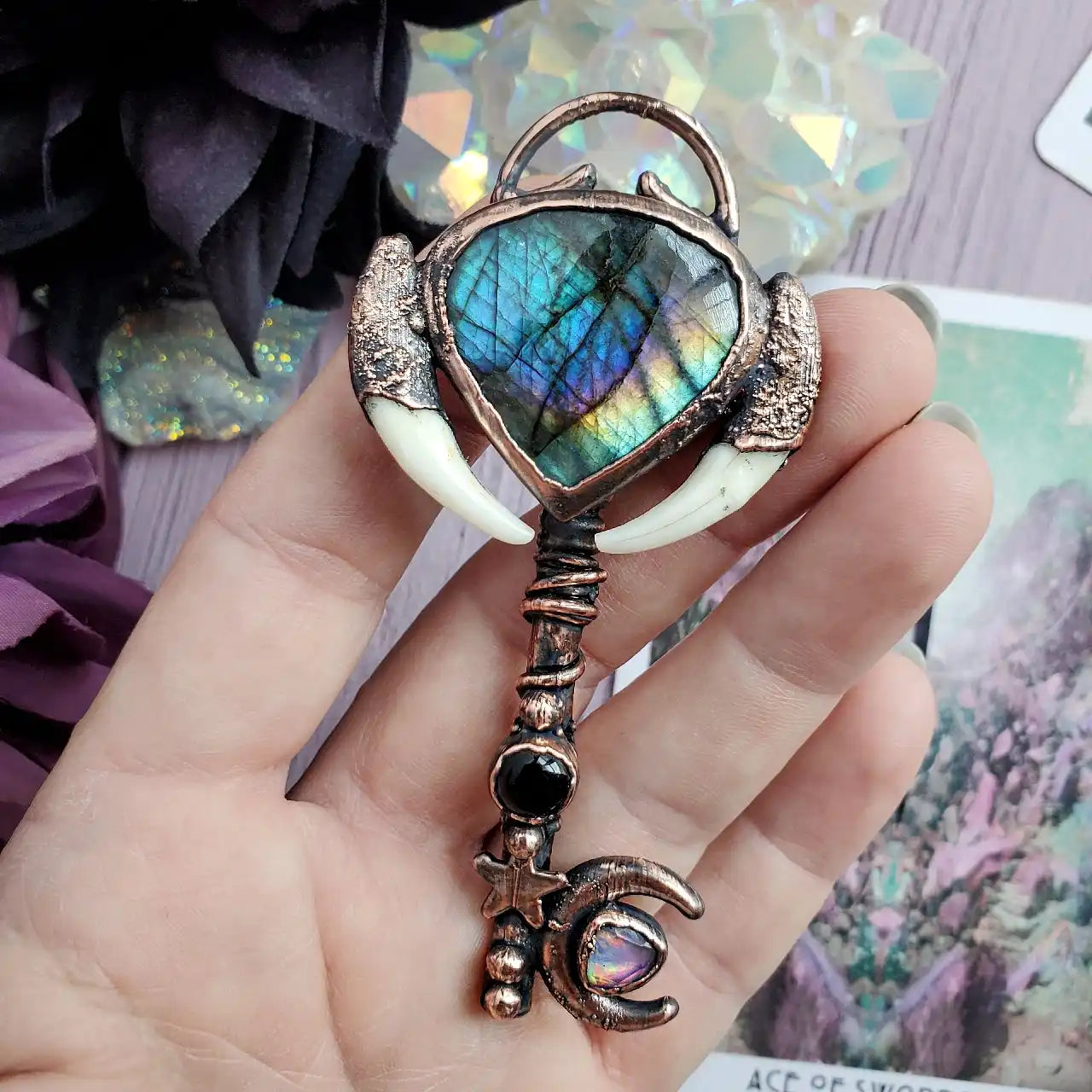 A copper key with a heart-shaped labradorite gemstone, two white fangs, a black gemstone, a star, and a crescent moon with an opal.