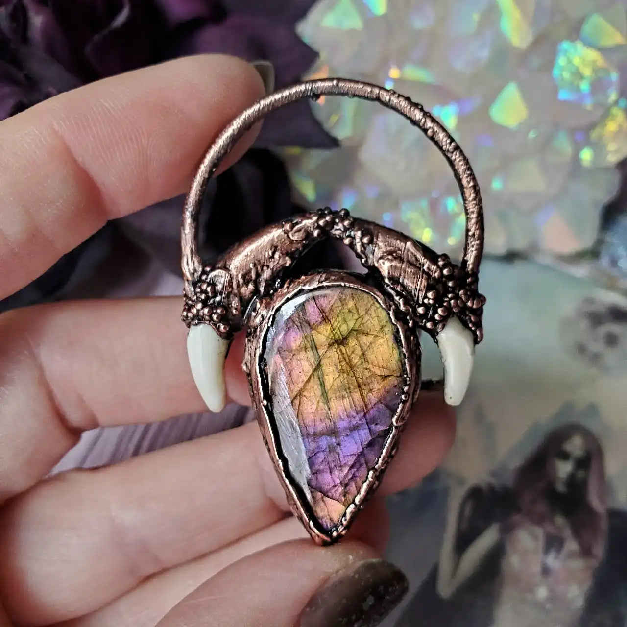A copper pendant featuring a tear-drop shaped labradorite stone with purple and yellow iridescence, flanked by two small white fangs.