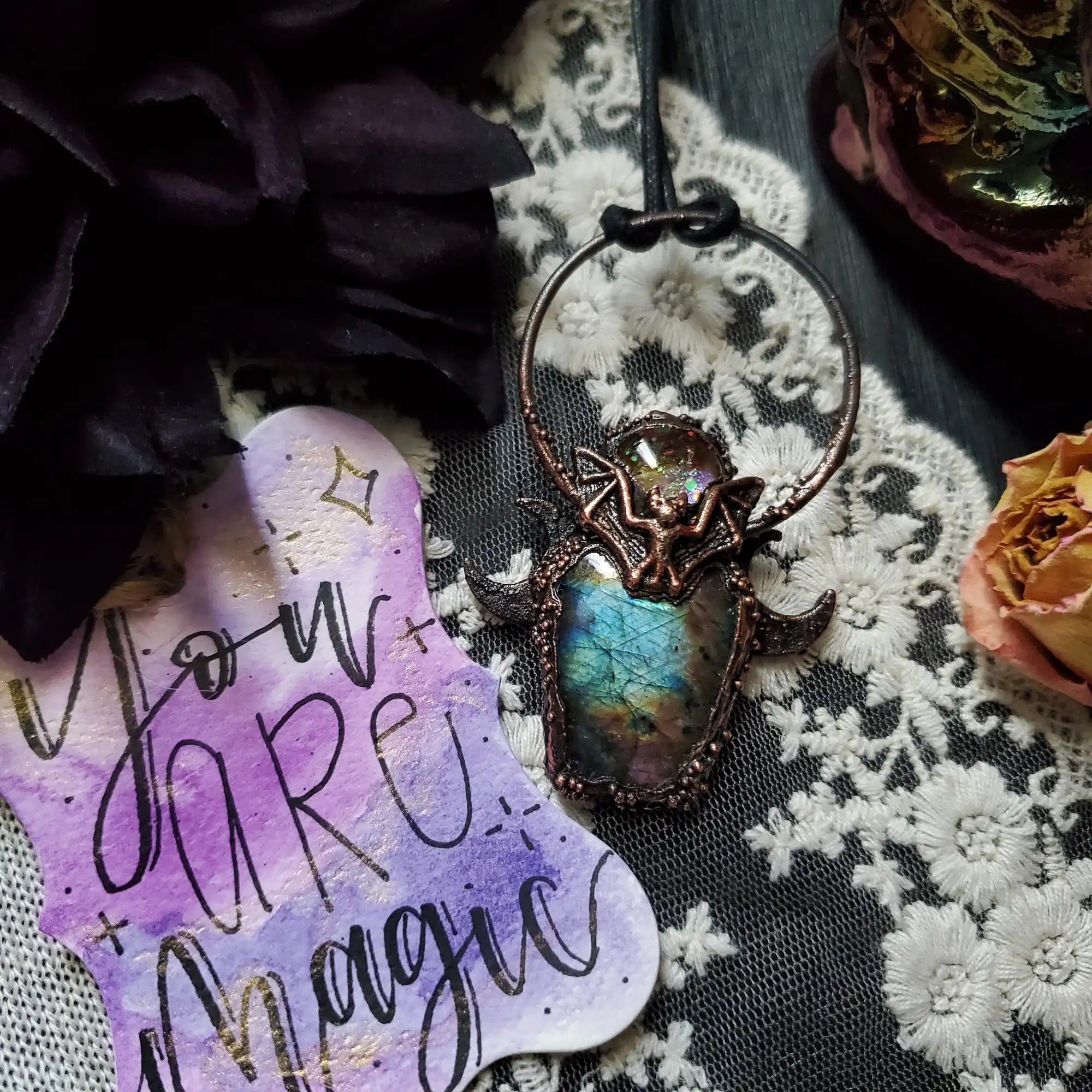 A copper-toned pendant featuring a central iridescent labradorite stone and a bat-like figure.