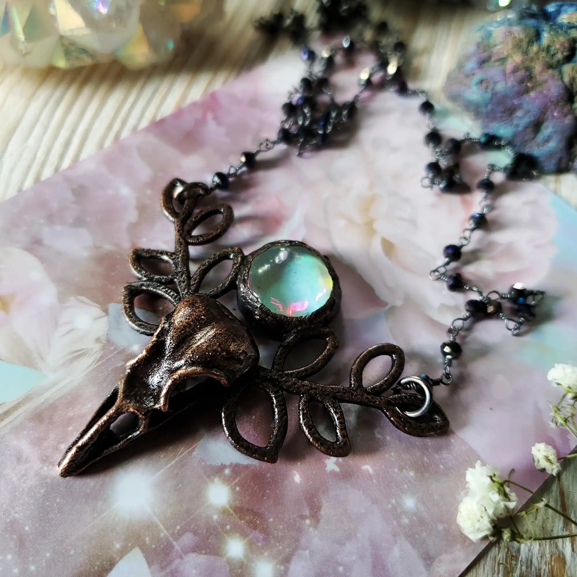 A dark bronze-colored necklace with a bird skull and leaf-like accents, featuring a central iridescent, opalescent gem.