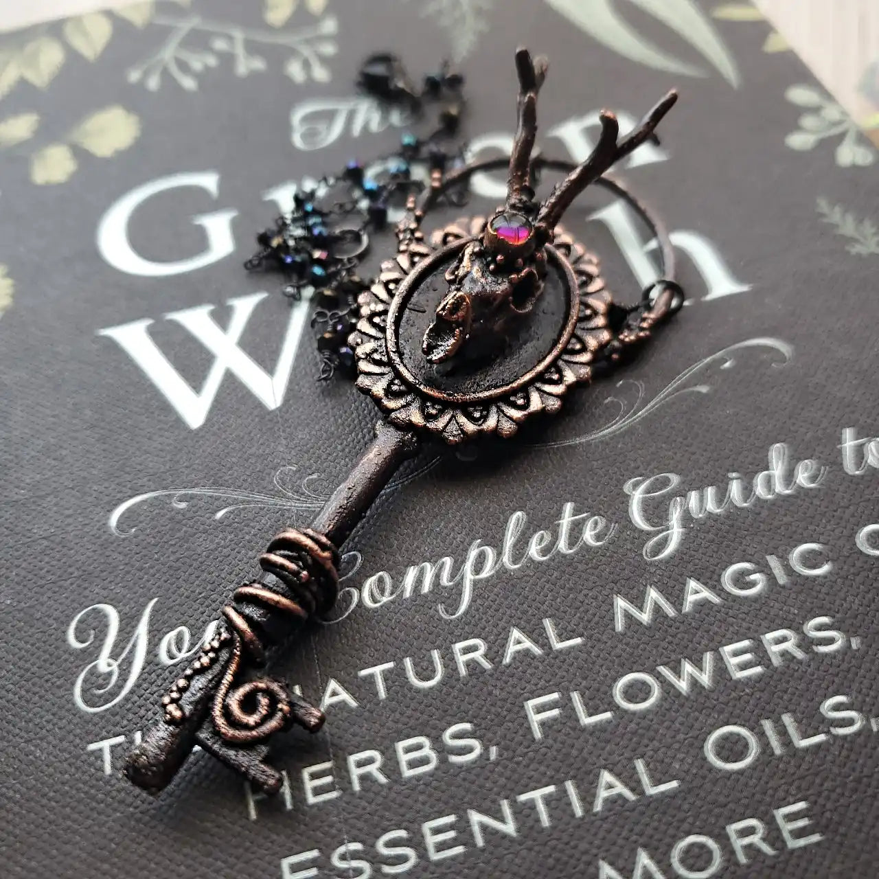 A dark bronze key-shaped pendant featuring a small animal skull with antlers and a shimmering iridescent gem.