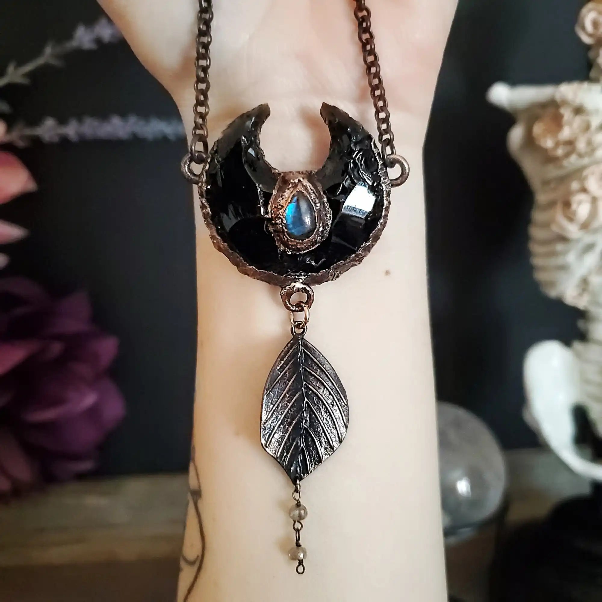 A dark crescent moon necklace featuring a blue gemstone and a dangling leaf charm with small beads.