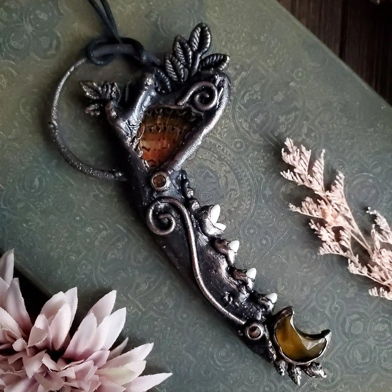 A dark metal fantasy-style pendant with leaf and swirl details, featuring a reddish-orange stone inlay and a yellow crescent moon-shaped stone at the tip.
