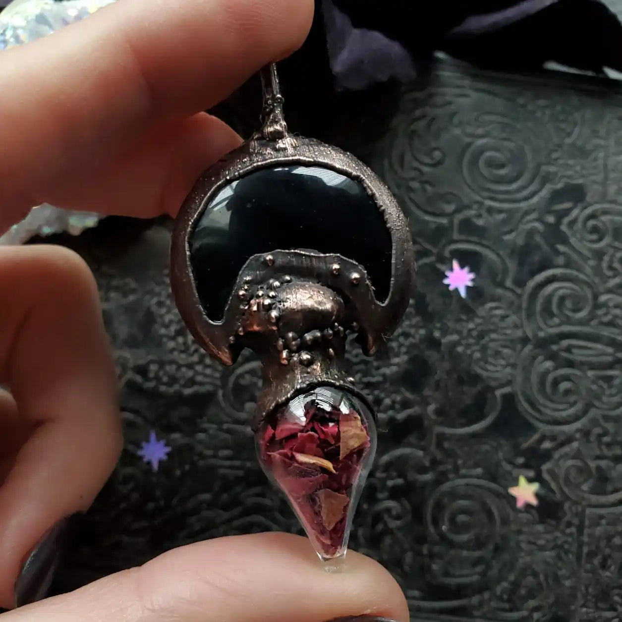 A dark, ornate metal pendant with a smooth black cabochon and a glass vial containing dried red petals.