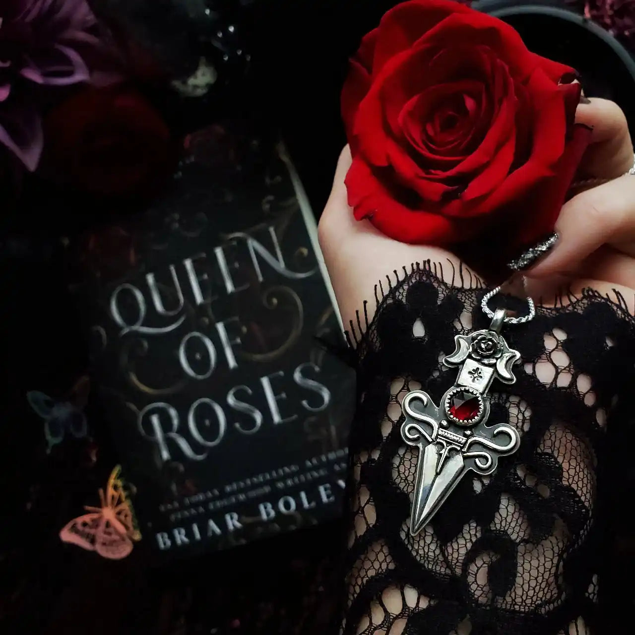 A hand adorned with a black lace cuff, holding a vibrant red rose and a silver dagger-shaped pendant with a red gem.