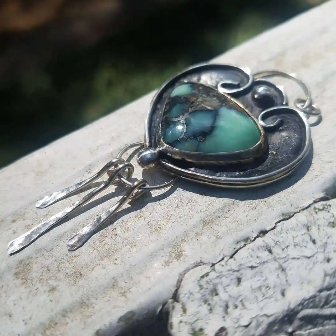 A handcrafted silver pendant featuring a vibrant green and black matrix turquoise gemstone, with three dangling silver pieces.