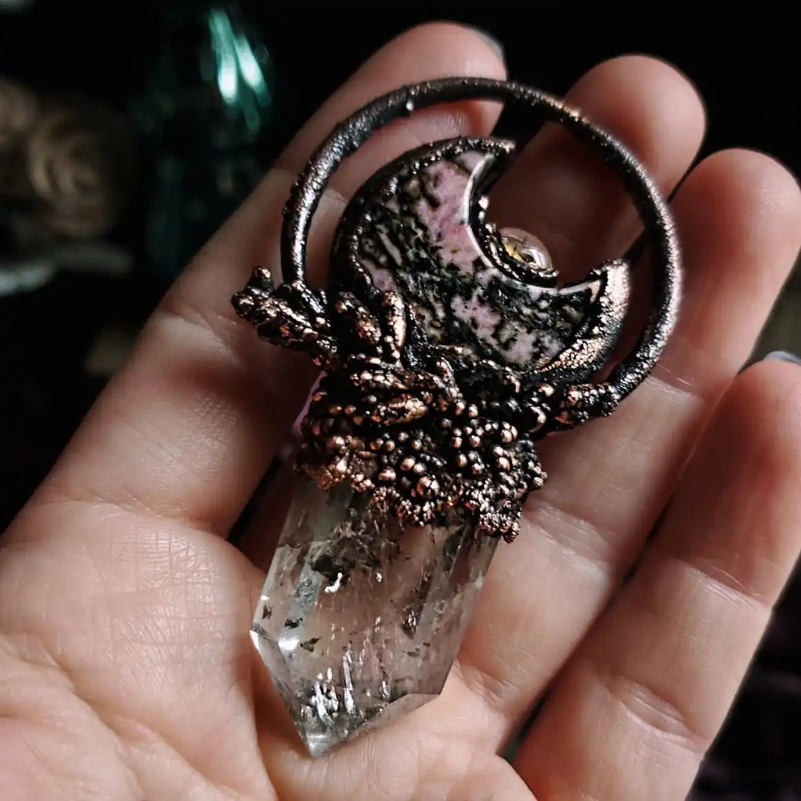 A handmade copper and crystal pendant featuring a crescent moon design with a pink and black stone inlay and a clear quartz point.