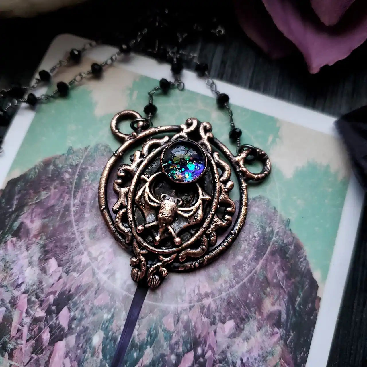 An ornate, dark metal necklace pendant featuring a bat design and a colorful, glitter-filled cabochon.