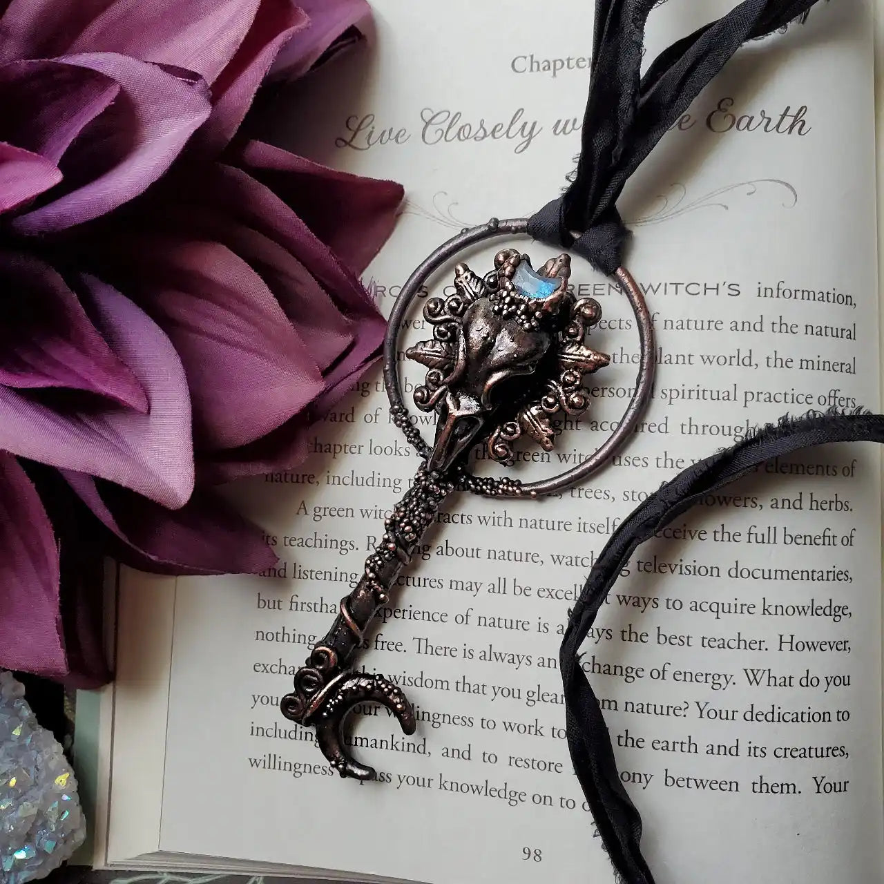 An ornate metal key featuring a crescent moon, a sunburst design, and a blue-hued gemstone, attached to a black fabric ribbon.