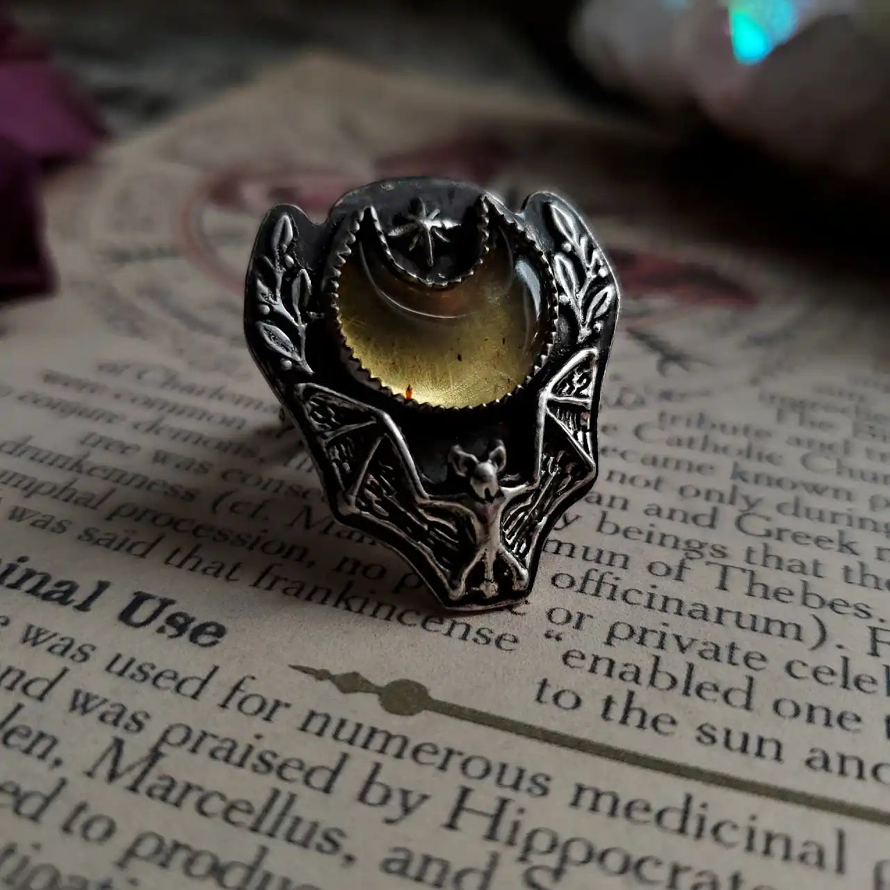 An ornate silver ring featuring a crescent moon with a star, a bat, and a yellowish-orange gemstone.