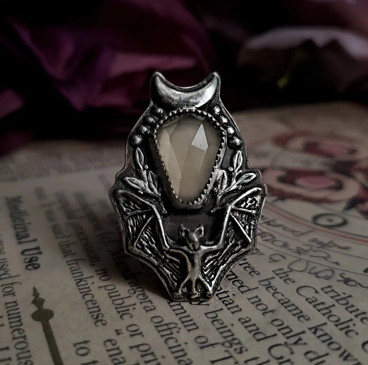 An ornate silver-toned ring with a central faceted off-white gemstone and a detailed bat motif.