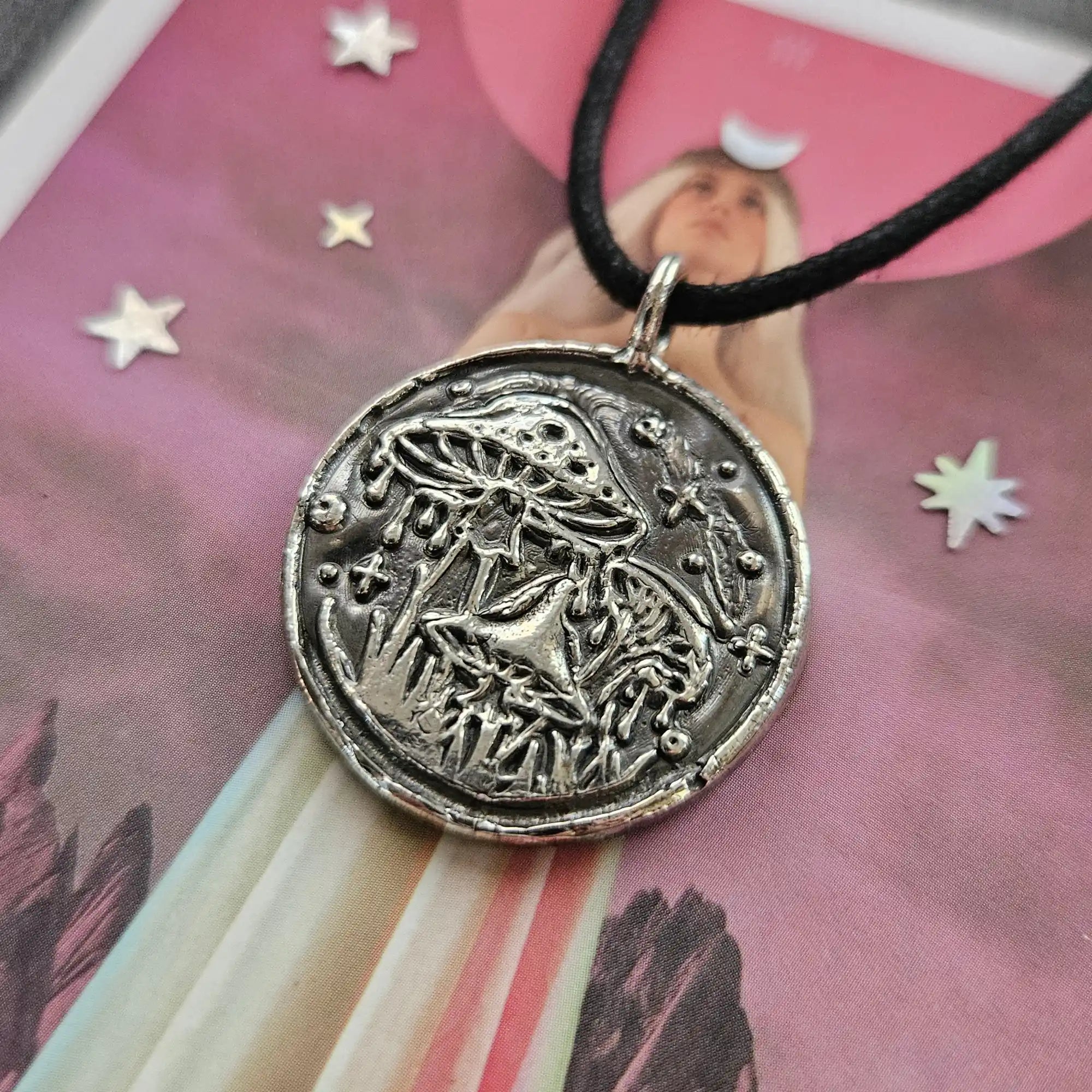 A round, silver-toned metal pendant with an embossed mushroom and celestial design on a black cord necklace.