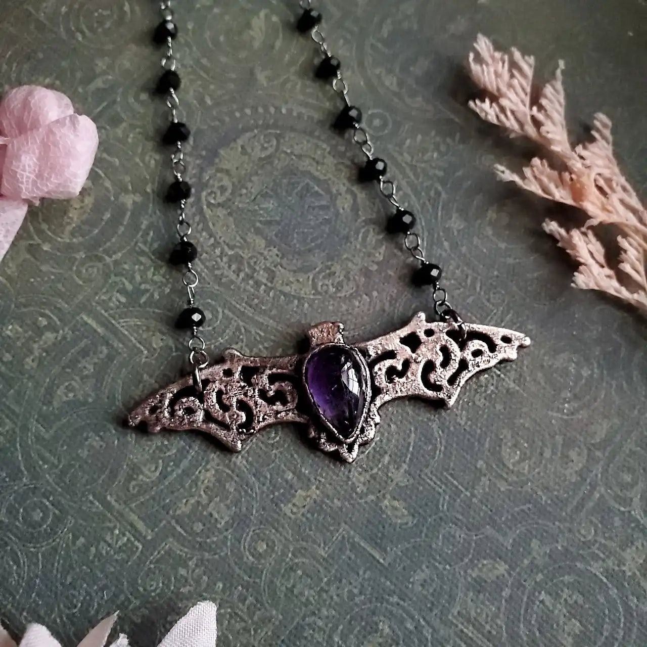 A silver-colored bat-shaped necklace pendant with a purple teardrop-shaped gemstone.