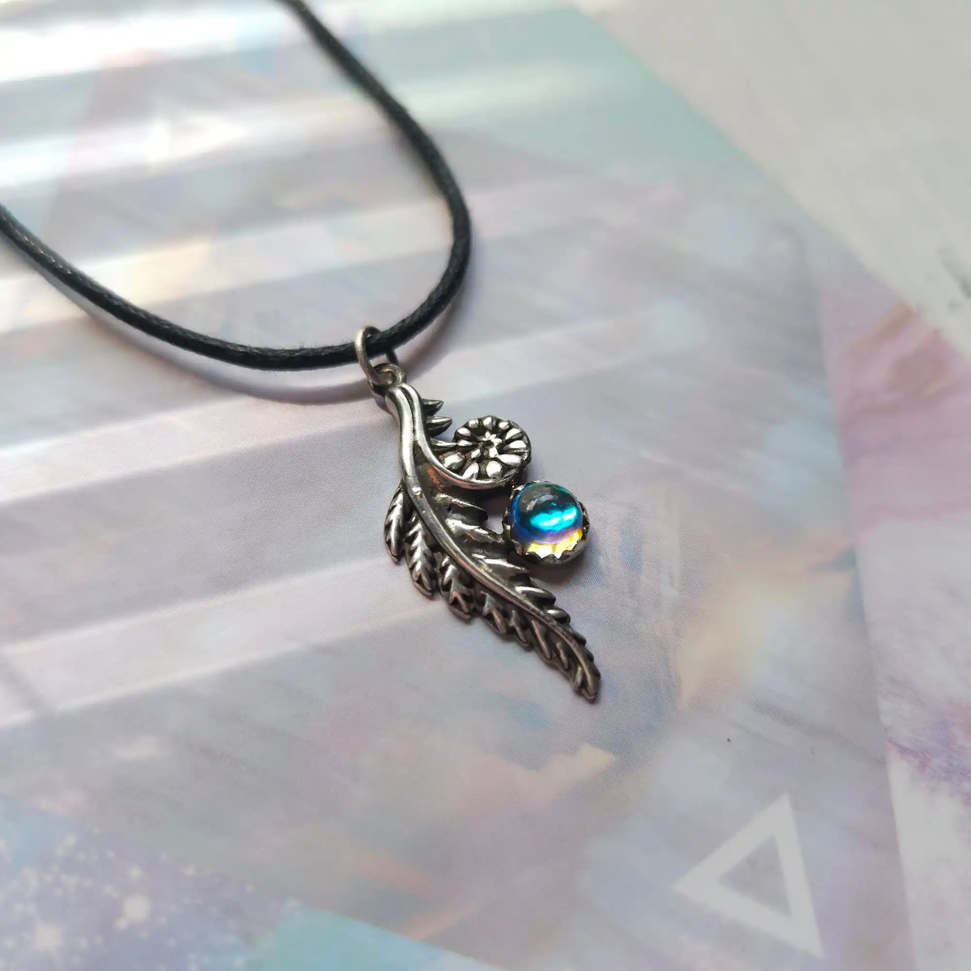 A silver-colored fern frond pendant with a blue-green iridescent gemstone, suspended from a black cord.