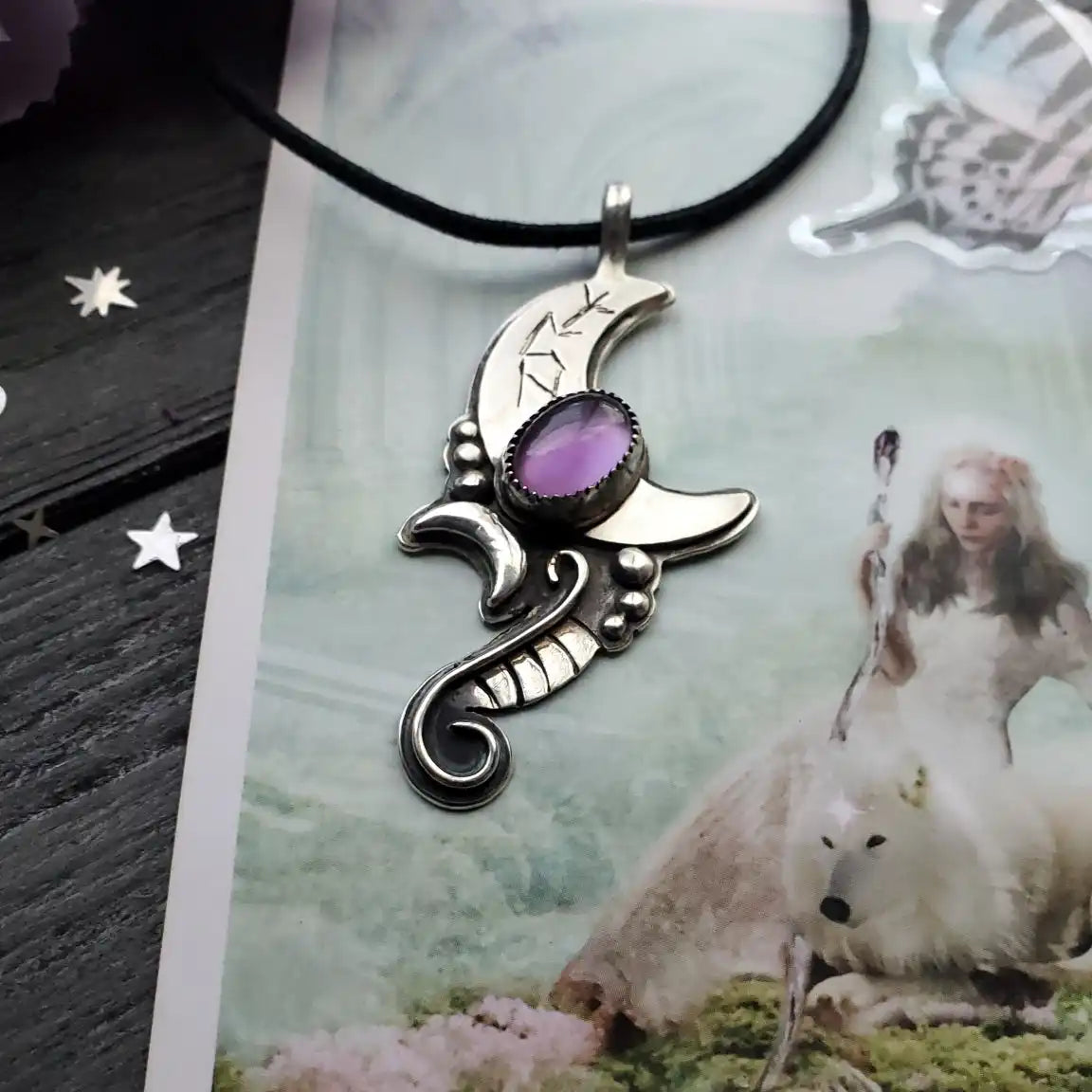 A silver crescent moon pendant with a central oval purple gemstone, featuring ornate swirling designs and etched symbols.