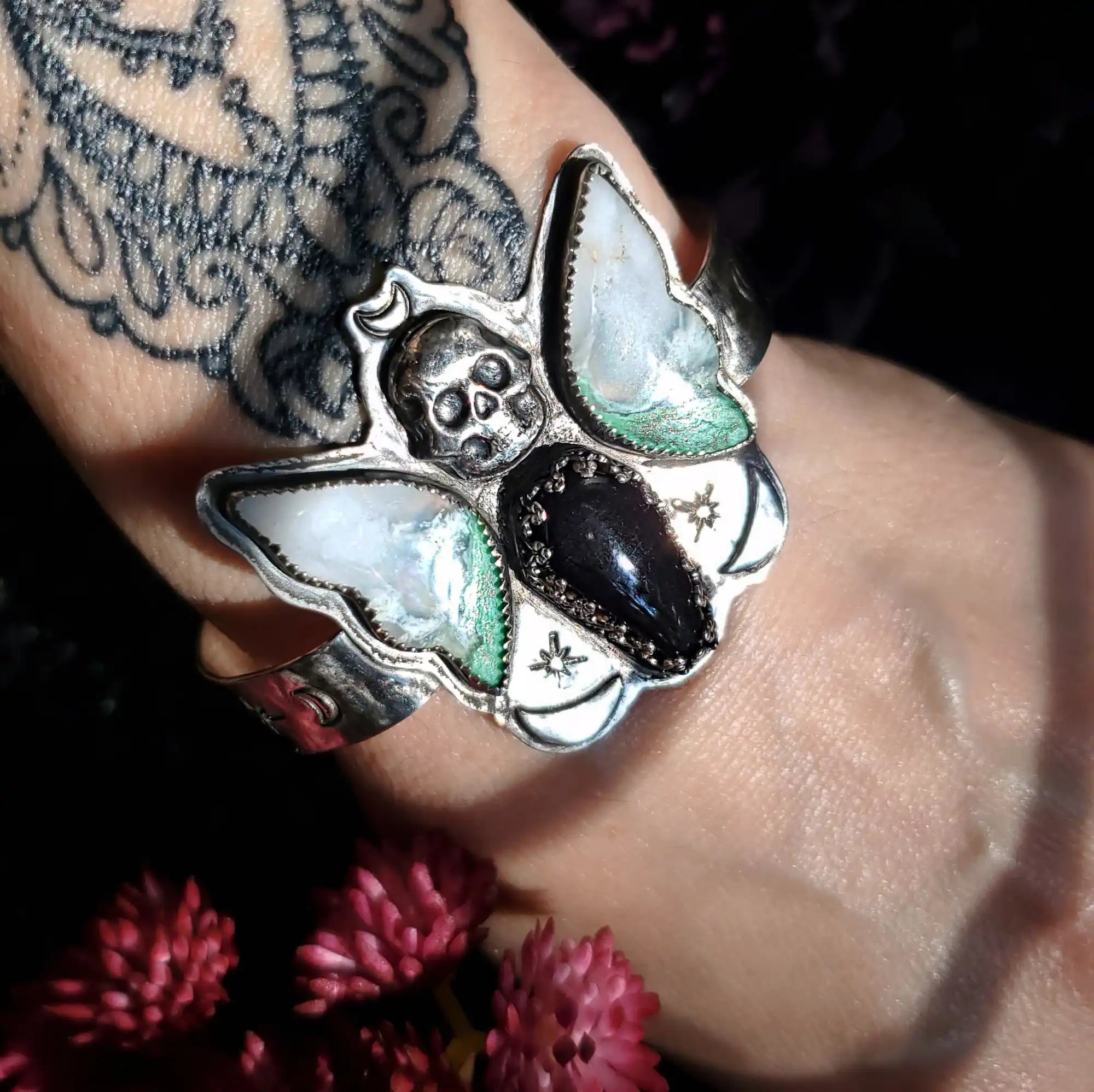A silver cuff bracelet adorned with a central skull, two pearlescent moth wings, and a black teardrop-shaped stone with a textured border, flanked by two small starburst designs.
