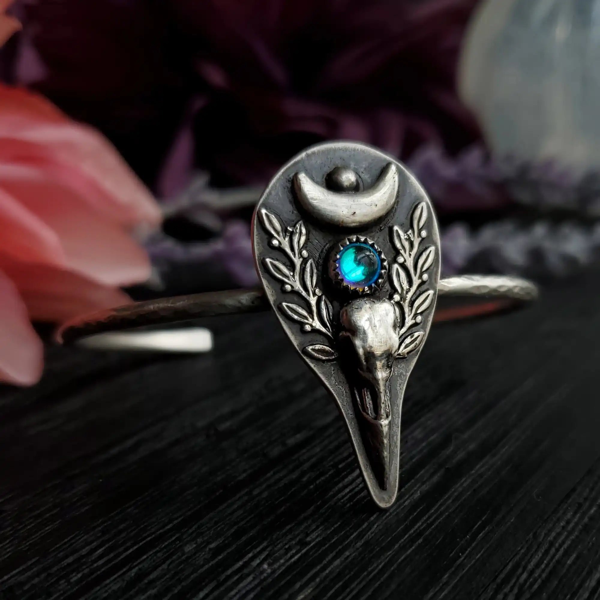 Silver cuff bracelet featuring a raven skull, crescent moon, and an iridescent blue gemstone.