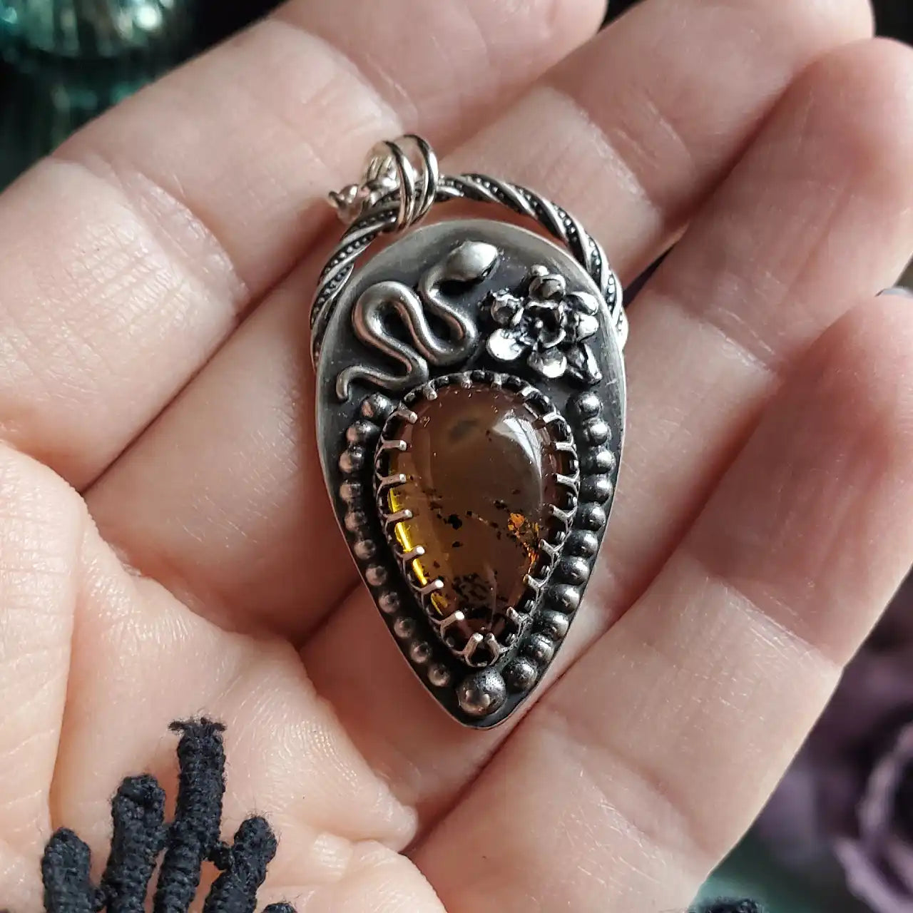 A silver pendant featuring a snake, a flower, and a teardrop-shaped amber stone with inclusions.