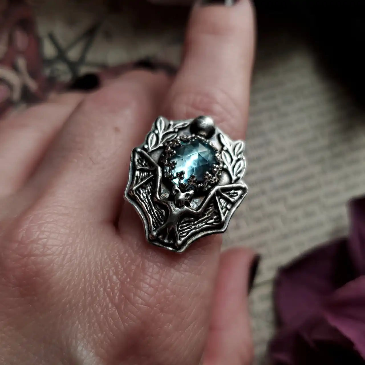 Silver ring with a central blue gemstone, flanked by a bat design and laurel wreaths.