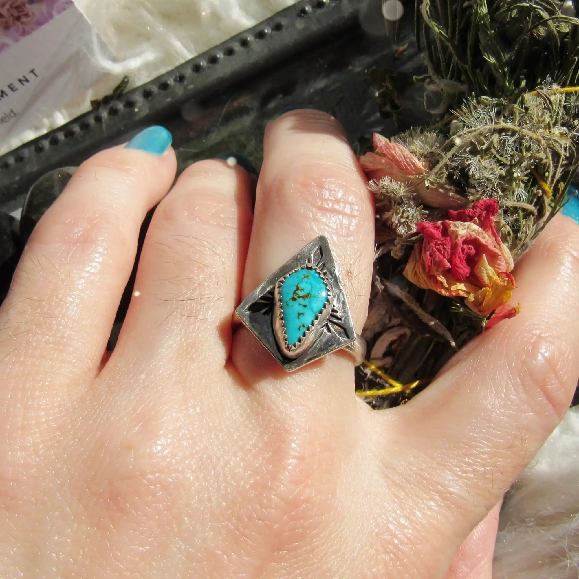 A silver ring with an engraved diamond-shaped setting holding a teardrop-shaped turquoise stone.