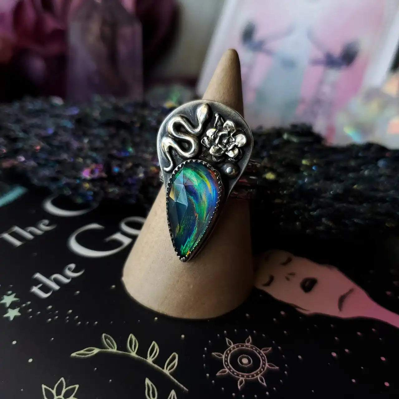 A silver ring featuring a winding snake and a floral design, with a faceted, teardrop-shaped iridescent gem.