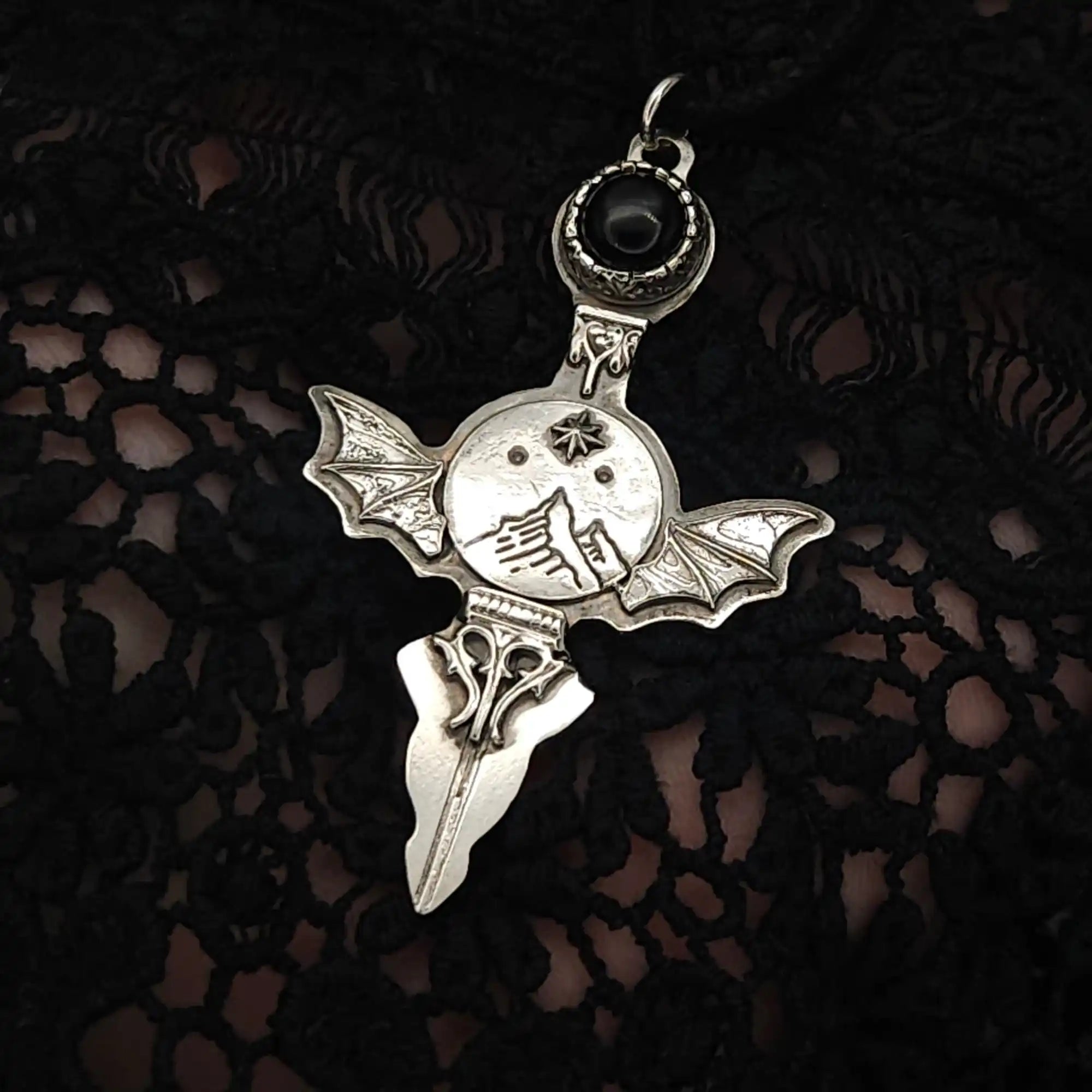 A silver-toned pendant featuring a bat-winged design with a central mountain motif and a dark stone at the top.