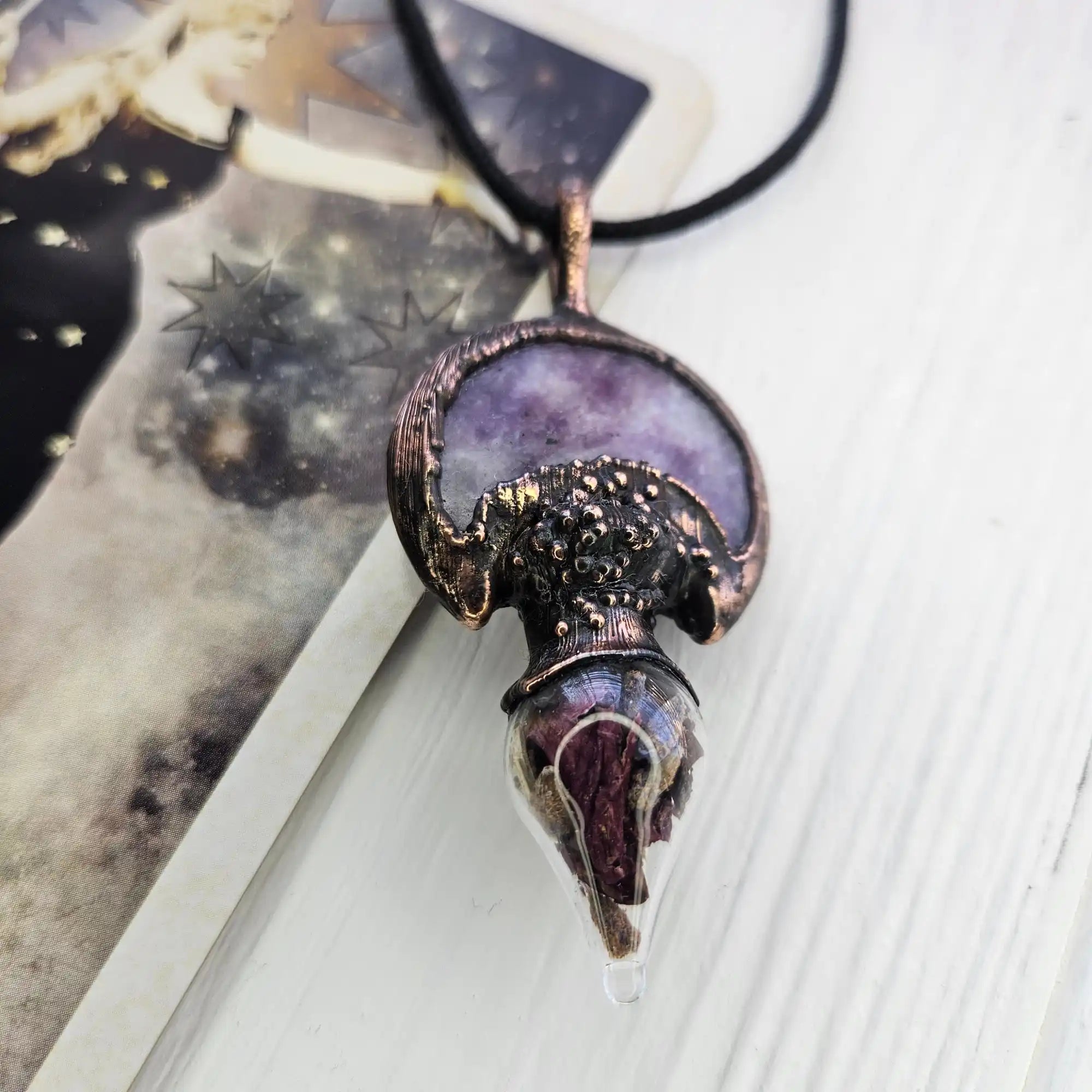 A unique, handcrafted pendant featuring a purple stone set in textured dark metal, with a clear, teardrop-shaped glass element below.