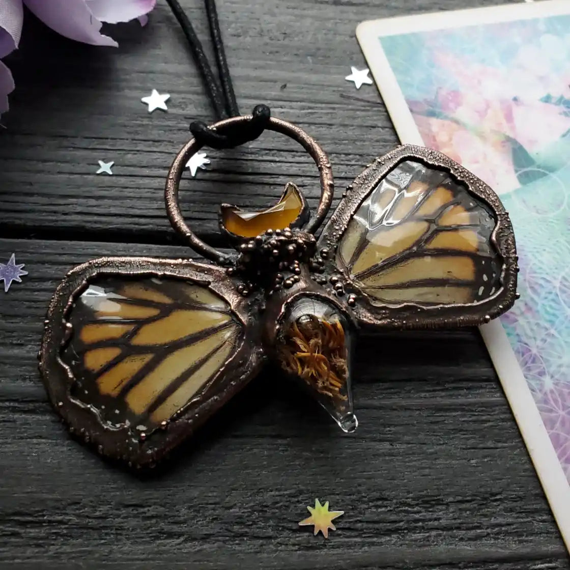 A unique necklace featuring real butterfly wings encased in copper, with an amber crescent moon accent and a glass drop containing natural fibers.